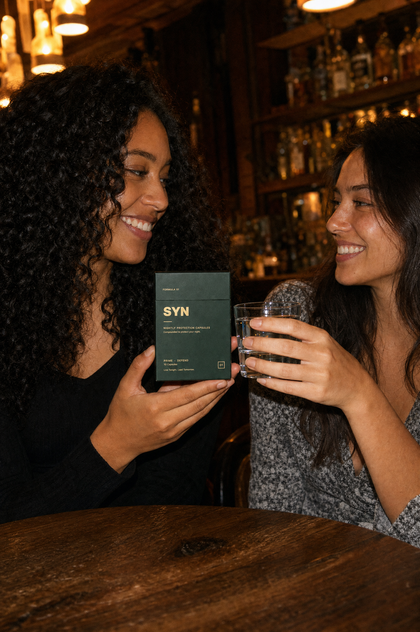 Two friends at a bar, one holding a box of SYN, one a glass of water