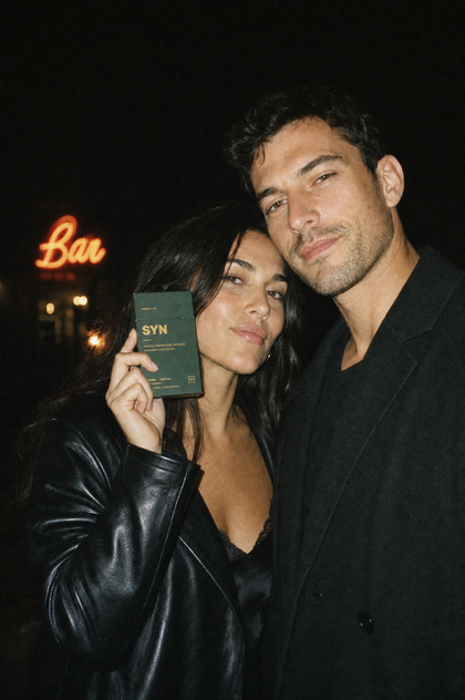 A couple outside a bar at night, holding a box of SYN