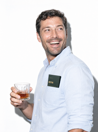A man laughing with a whisky, a box of SYN in his shirt pocket